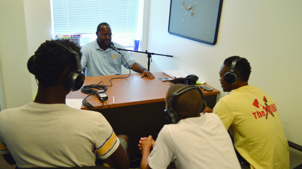 Three youth wearing headphones interviewing an adult speaking into a microphone.