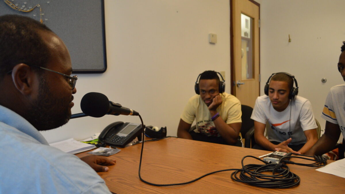 Three youth wearing headphones interviewing an adult speaking into a microphone.