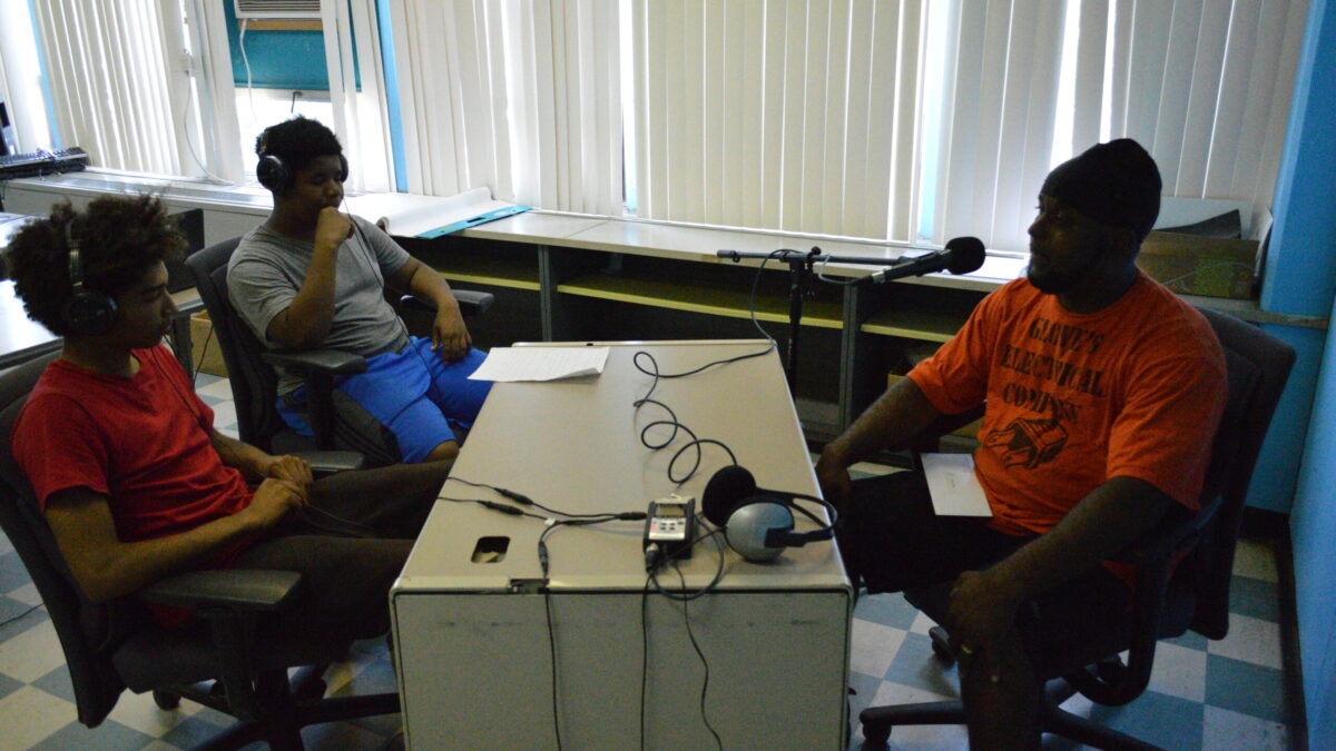 Two youth wearing headphones interviewing an adult speaking into a microphone