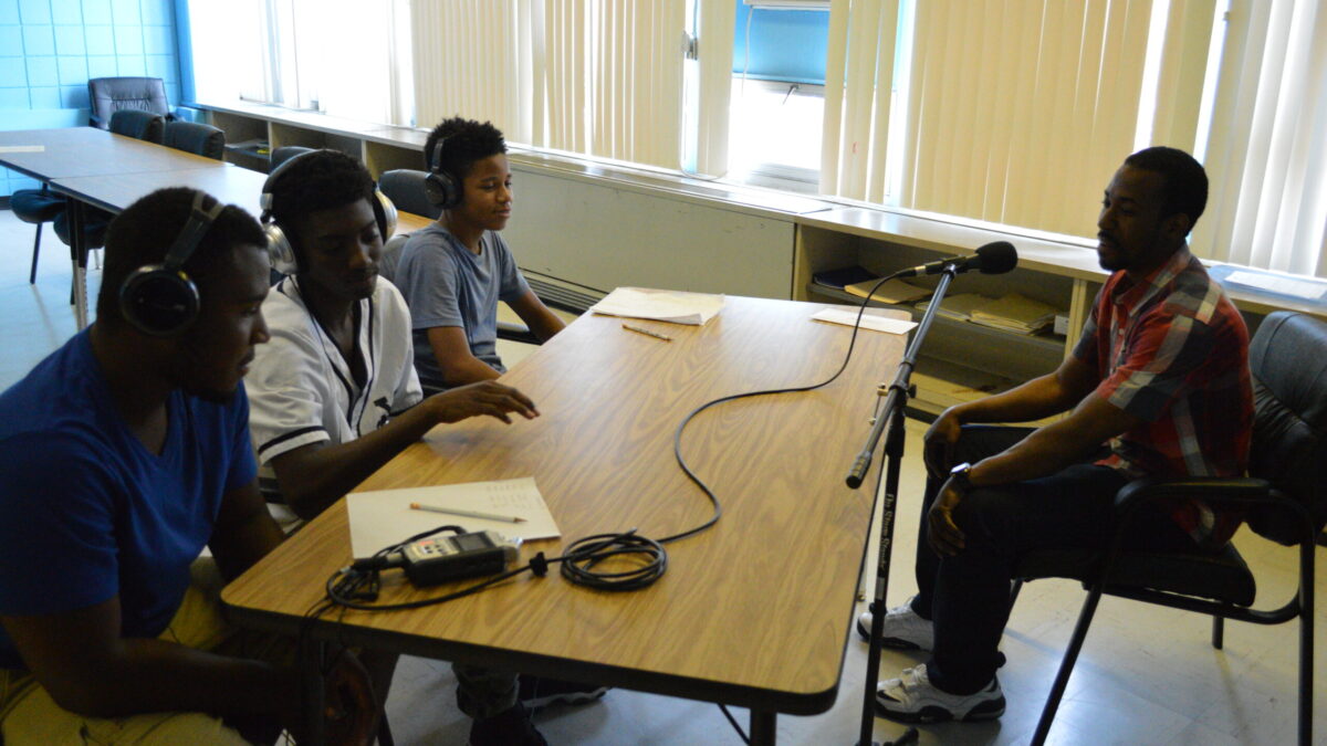 Three youth wearing headphones interviewing an adult speaking into a microphone