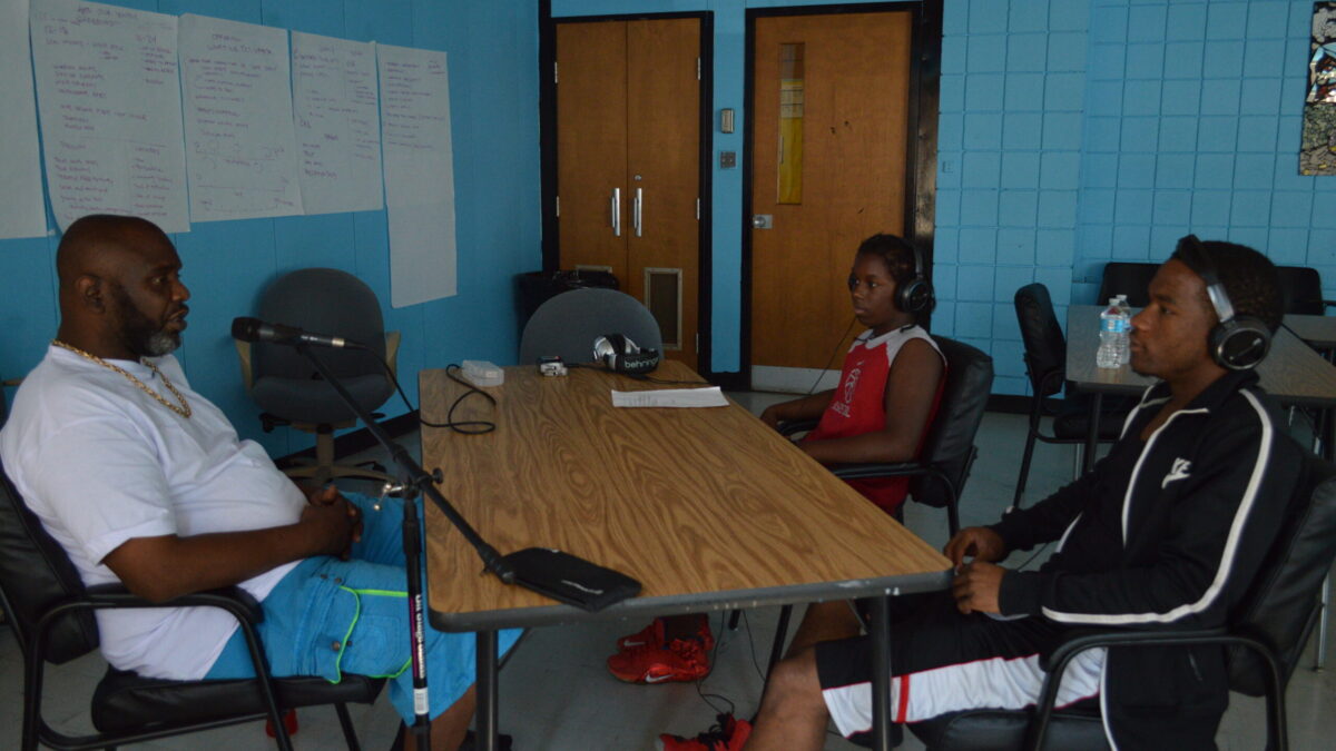 Two youth wearing headphones interviewing an adult speaking into a microphone