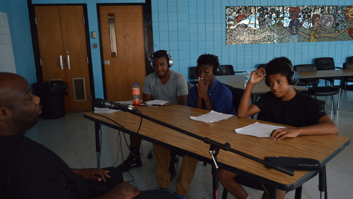 Three youth wearing headphones interviewing an adult speaking into a microphone
