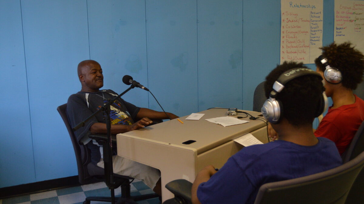 Two youth wearing headphones interviewing an adult speaking into a microphone