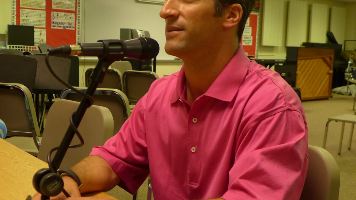 An adult speaking into a microphone sitting at a long wooden table