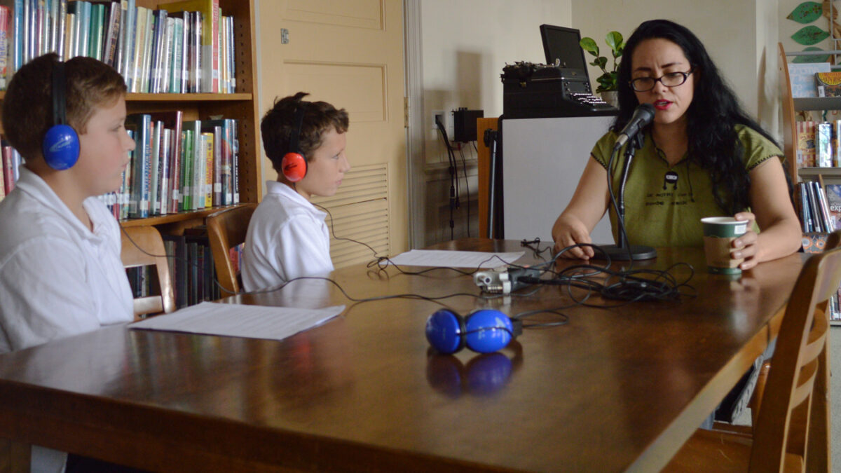 Two youth wearing headphones interviewing an adult speaking into a microphone in a library