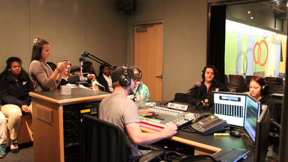 Three youth wearing headphones sitting behind microphones, five youth sitting off to the side, an adult filming on a smartphone, and an adult wearing headphones sitting behind a mixing board in a radio studio
