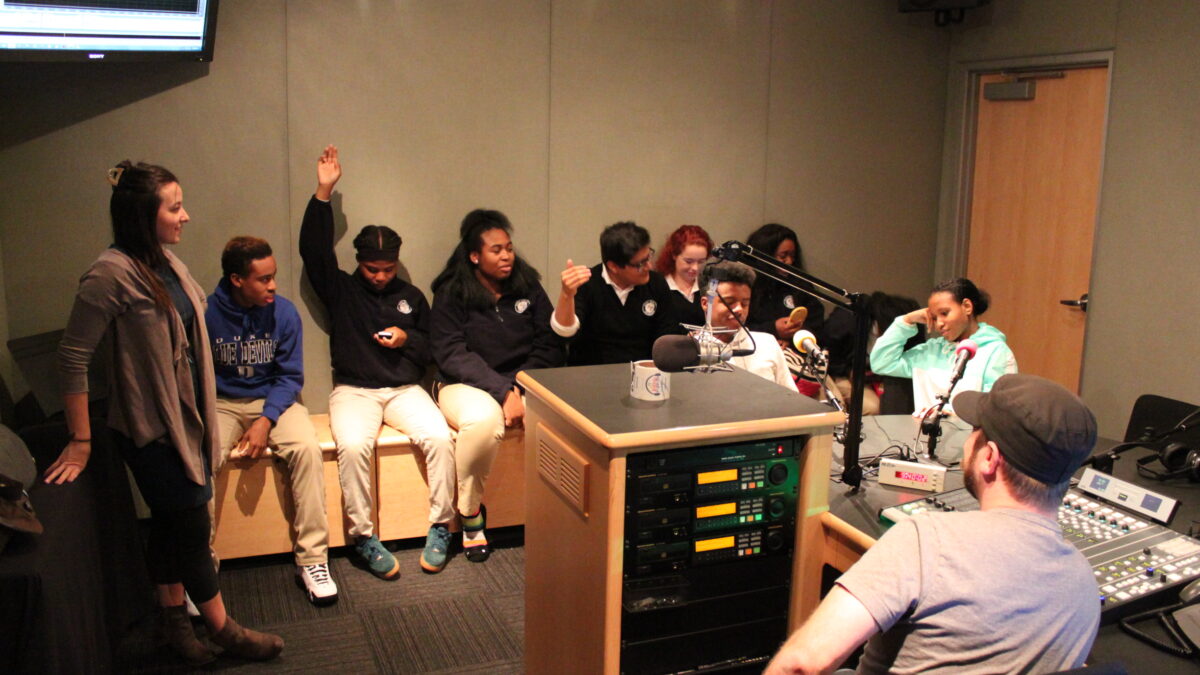 A youth sitting behind a microphone, six youth sitting off to the side, two raising their hands, an adult standing off to the side, and an adult sitting behind a mixing board looking at the youth in a radio studio