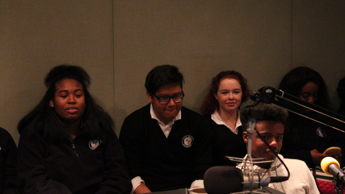 Four youth sitting off to the side and one youth sitting behind a microphone in a radio studio