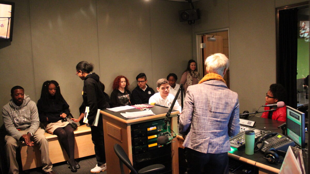 Seven youth sitting off to the side, two youth sitting behind microphones, and an adult standing in a radio studio