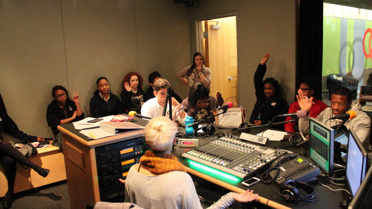 Four youth sitting behind microphones, one holding up their arm, four youth sitting off to the side, one holding up their hand, an adult standing off to the side, and an adult sitting behind a mixing board in a radio studio