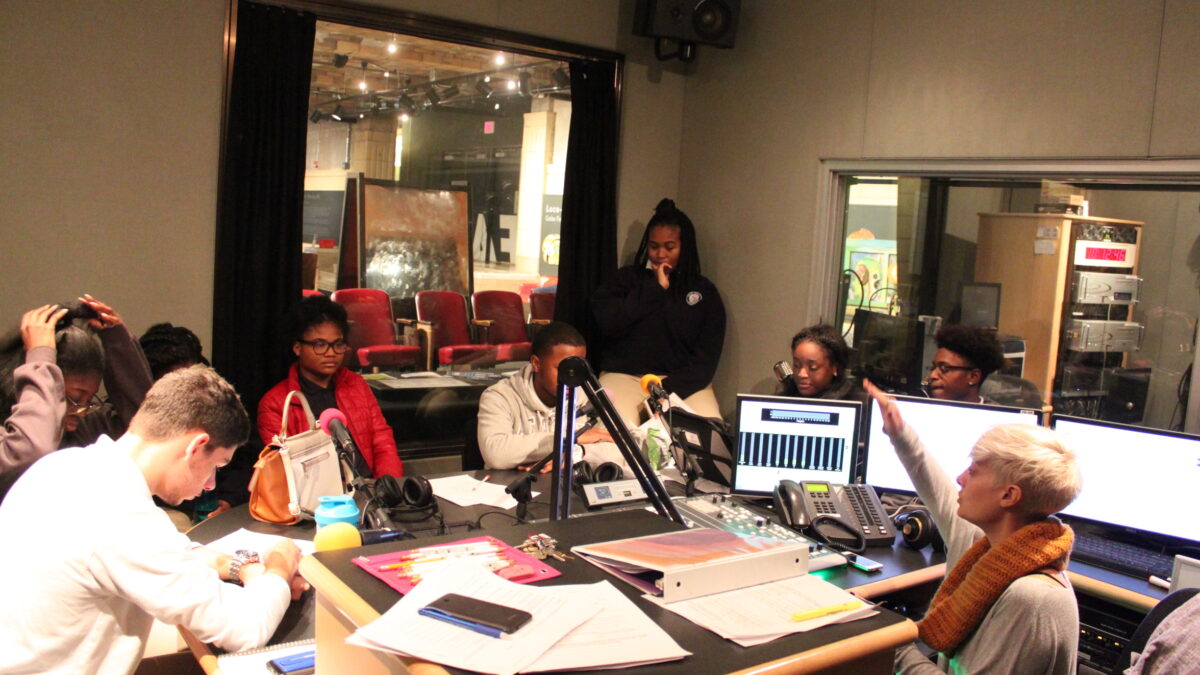 Six youth sitting behind microphones looking down at pieces of paper, one youth standing in the corner, and an adult sitting behind a mixing board in a radio studio