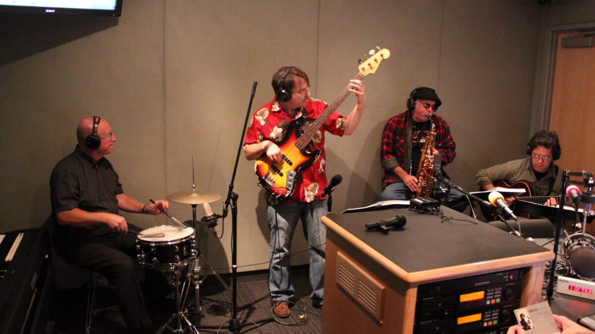 IMG_2099 An adult wearing headphones playing drum set, an adult wearing headphones playing an electric bass, an adult wearing headphones playing alto saxophone, and an adult wearing headphones playing electric guitar in a radio studio