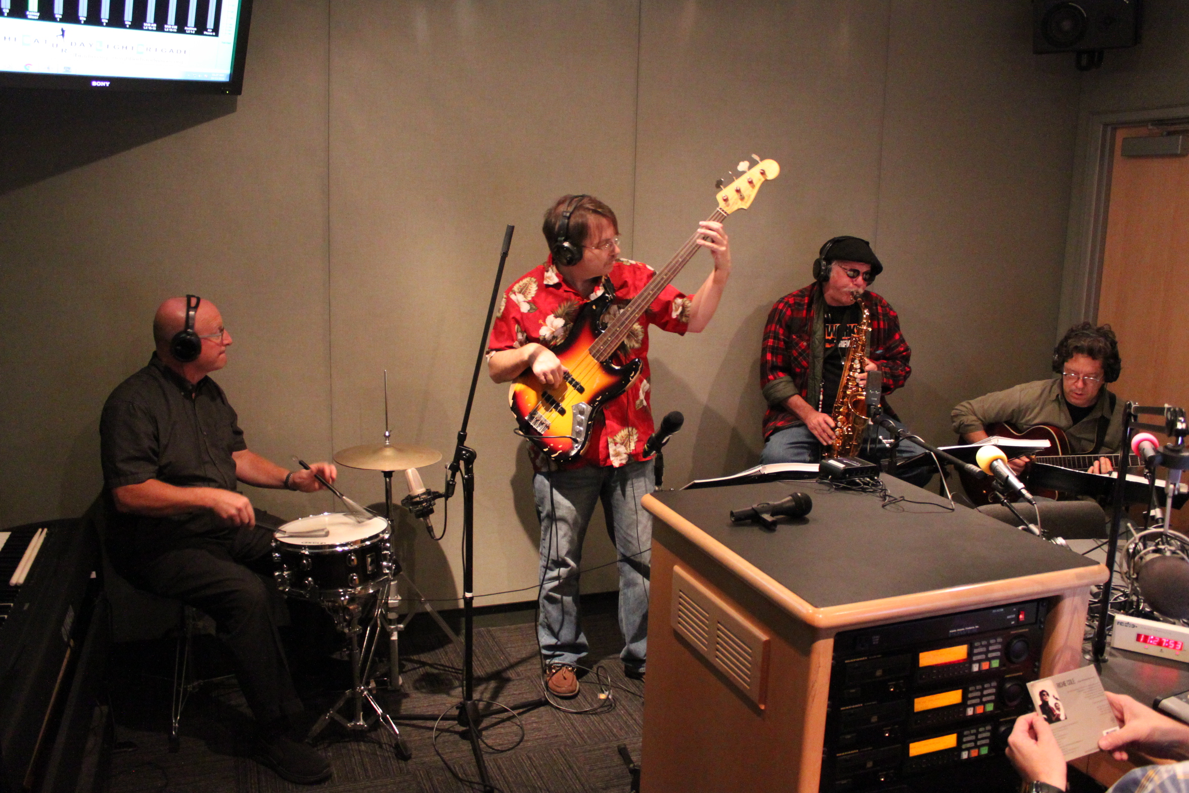 An adult wearing headphones playing drum set, an adult wearing headphones playing an electric bass, an adult wearing headphones playing alto saxophone, and an adult wearing headphones playing electric guitar in a radio studio