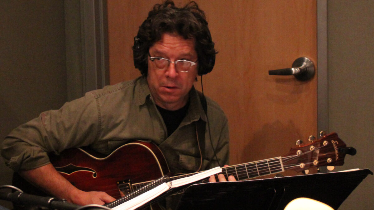 IMG_2102 An adult wearing headphones playing guitar in a radio studio