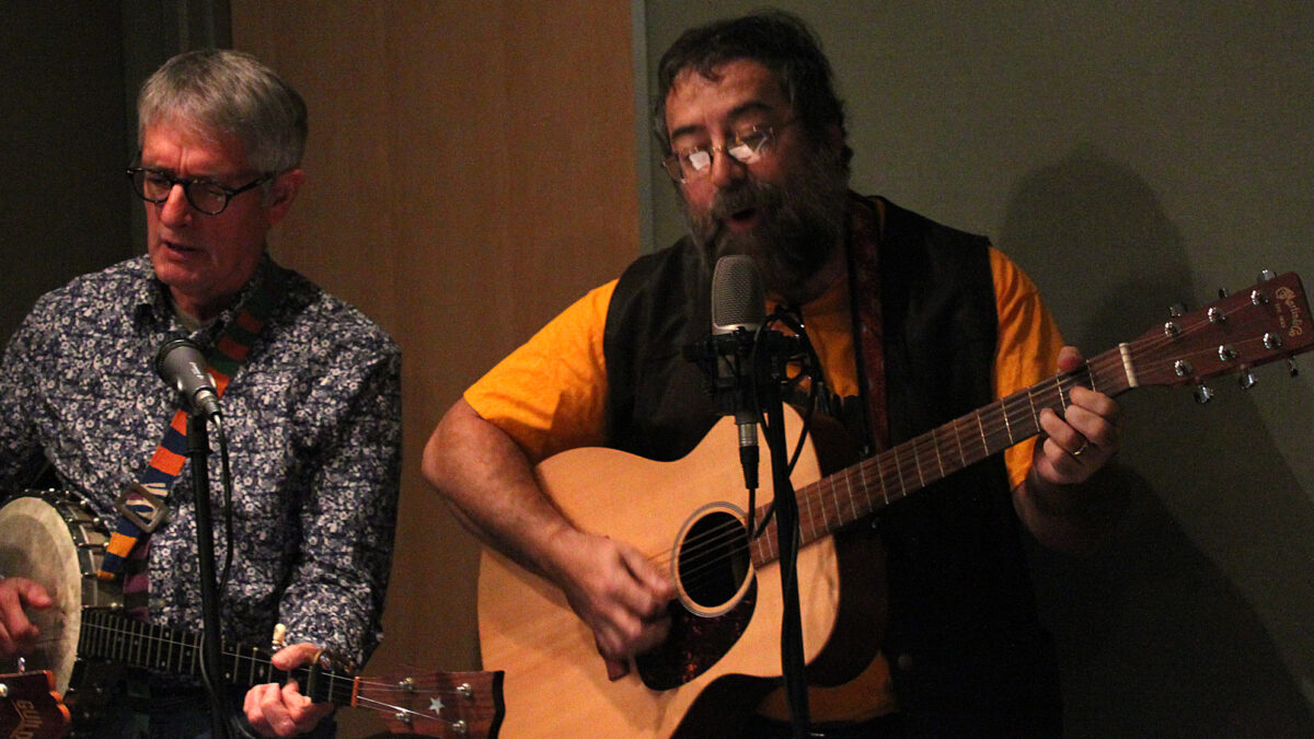 An adult playing a banjo and an adult playing an acoustic guitar, both singing into microphones in a radio studio
