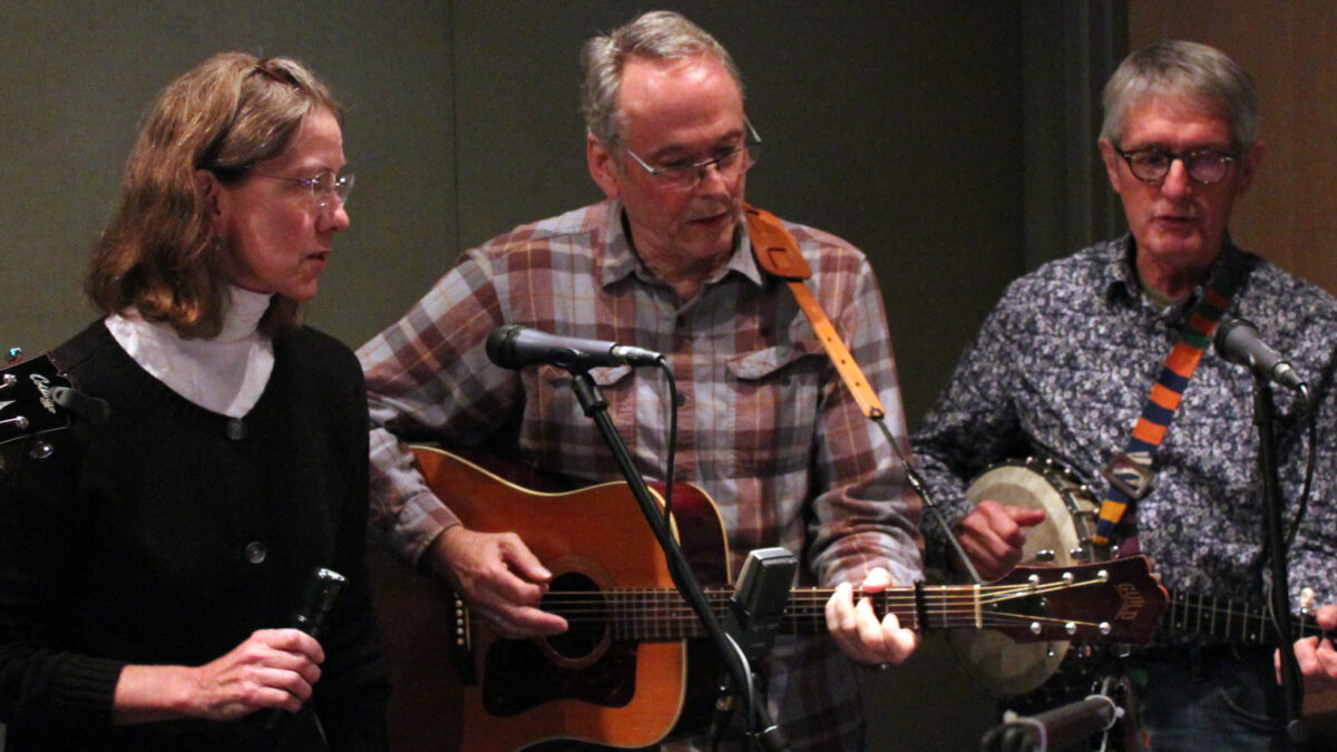 An adult holding a shaker and singing into a microphone, an adult playing an acoustic guitar, and an adult playing a banjo in a radio studio
