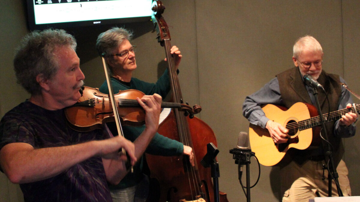 An adult playing a fiddle, an adult playing an upright bass, and an adult playing an acoustic guitar in a radio studio
