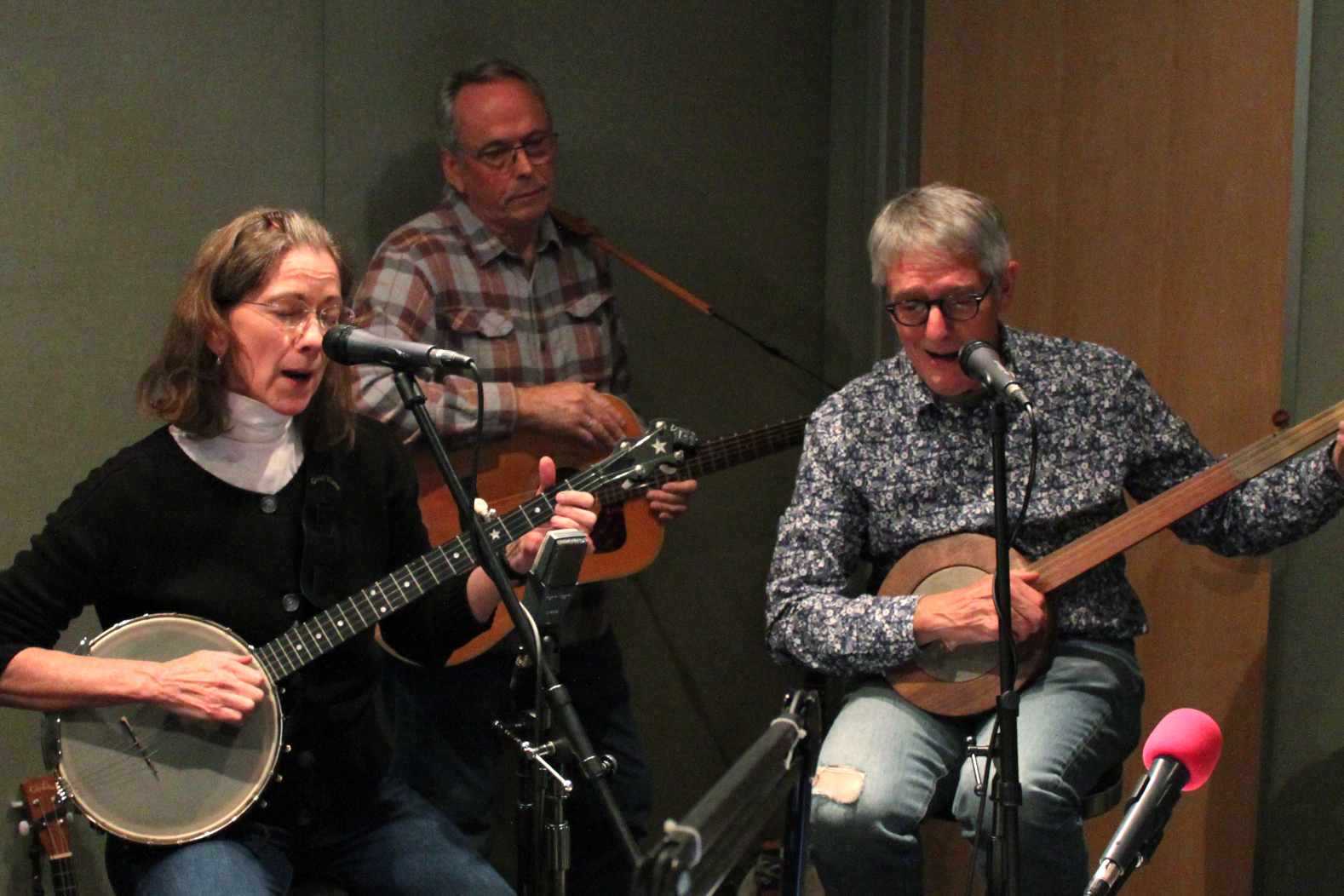 Two adults playing banjos and singing into microphones and an adult playing an acoustic guitar in a radio studio