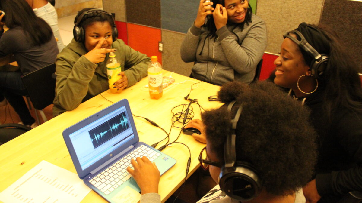 Four youth wearing headphones sitting around a laptop computer on a wooden table