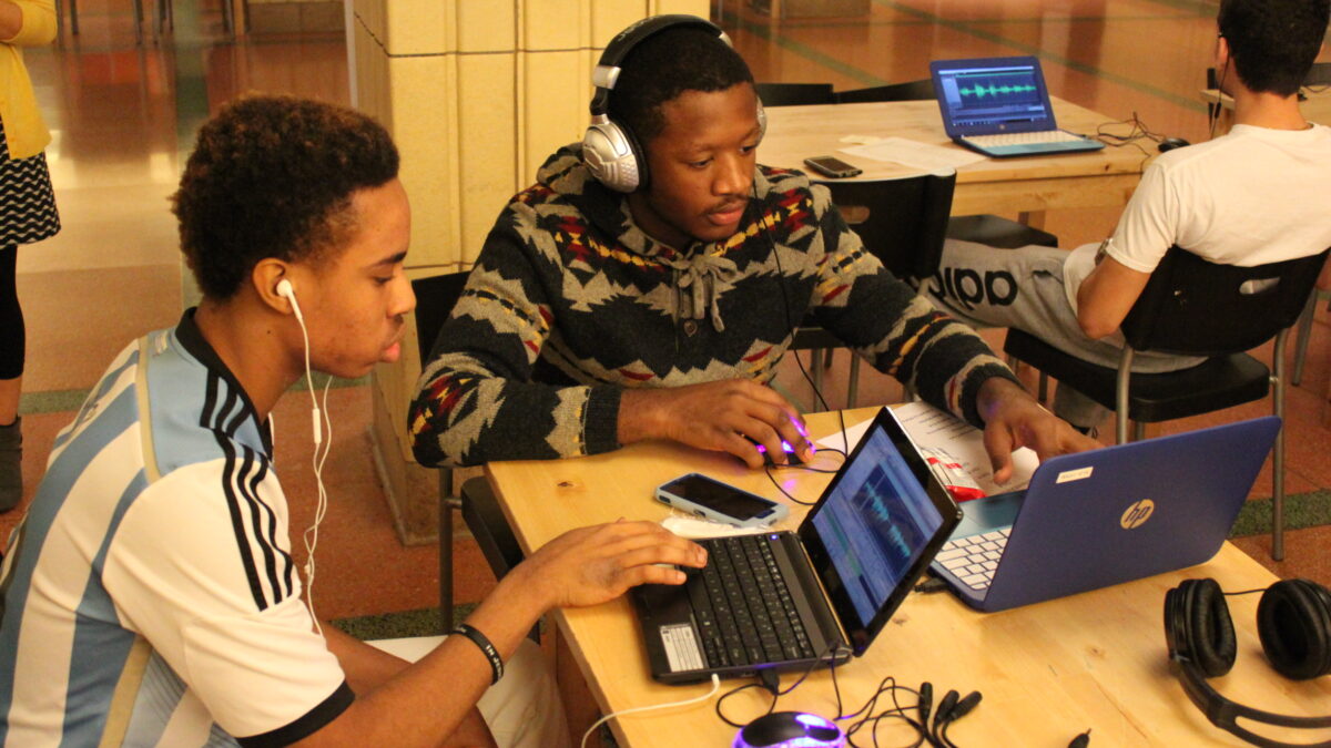 Two teens wearing headphones looking at laptop computers on a wooden table