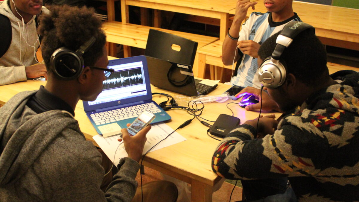 Four teens sitting around a laptop computer on a wooden table, two teens wearing headphones