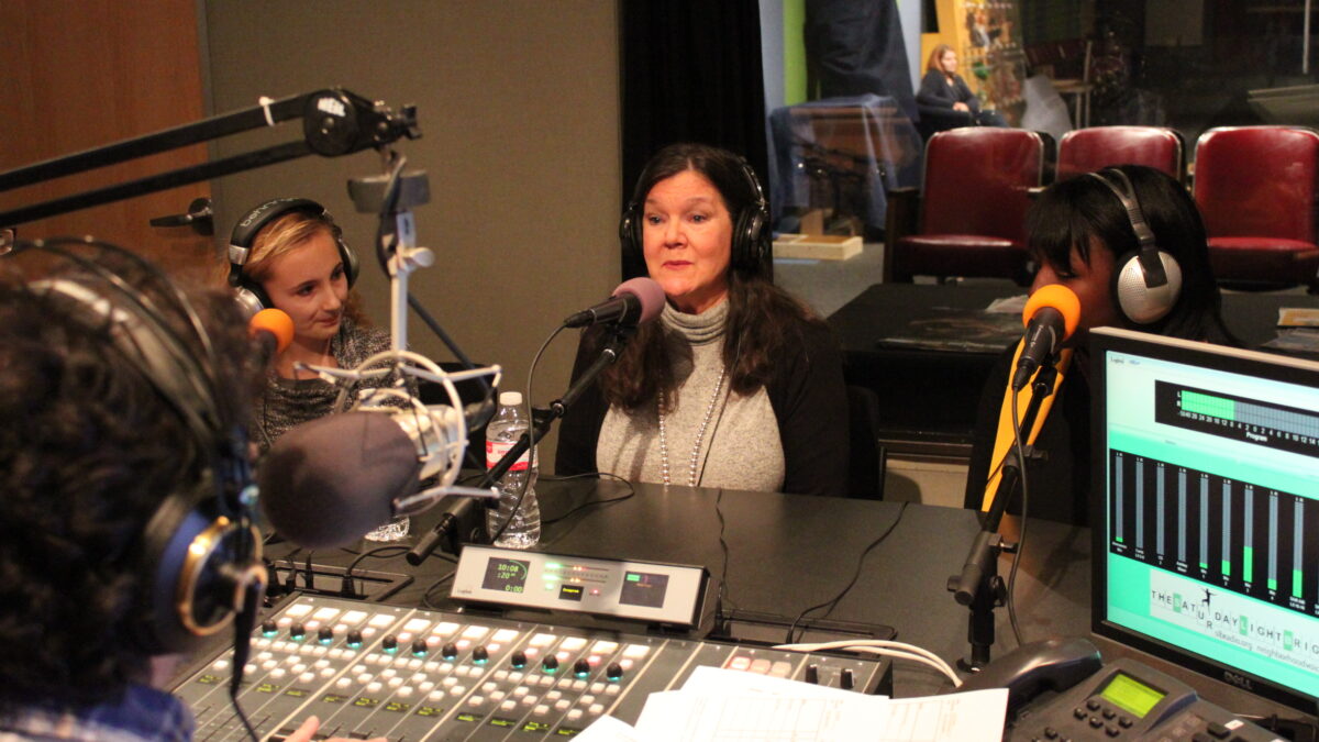 Three teens wearing headphones speaking into microphones being interviewed by an adult wearing headphones sitting behind a microphone and a mixing board in a radio studio