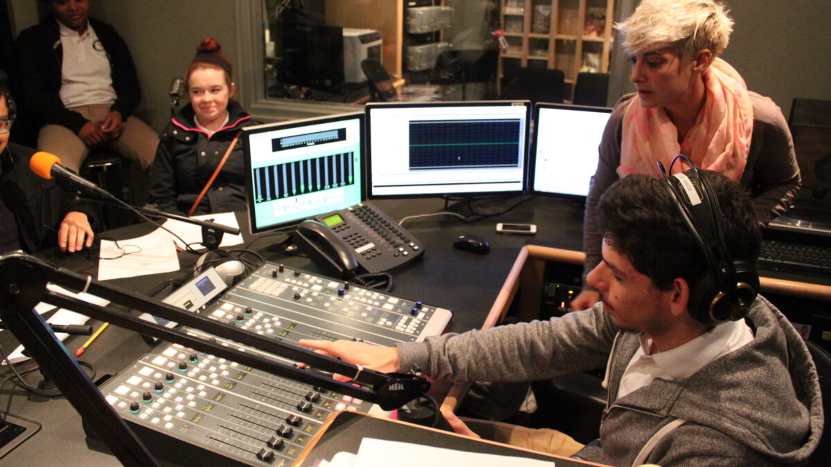 An adult looking at a teen wearing headphones interacting with a mixing board and two youth sitting in the background in a radio studio