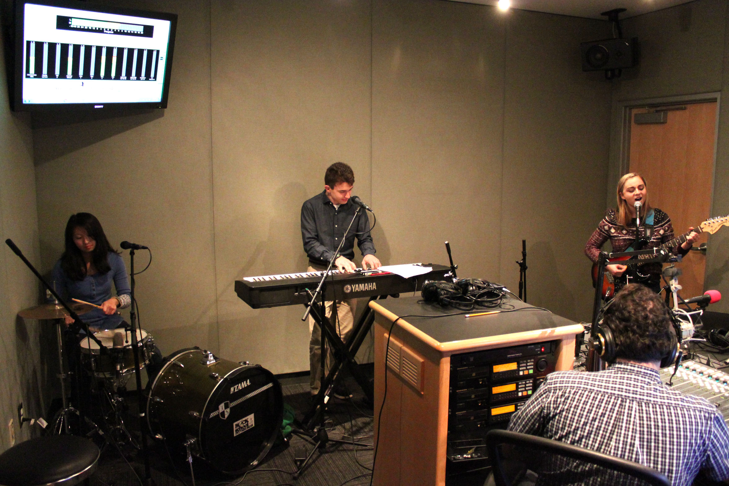 A teen playing a drum set, a teen playing a piano keyboard, and a teen playing electric guitar as an adult wearing headphones sitting behind a mixing board in a radio studio