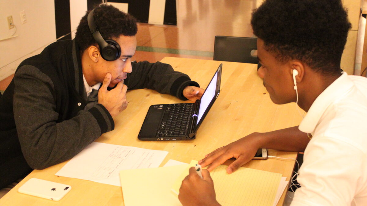 Two teens wearing headphones, one looking at a laptop computer on a wooden table