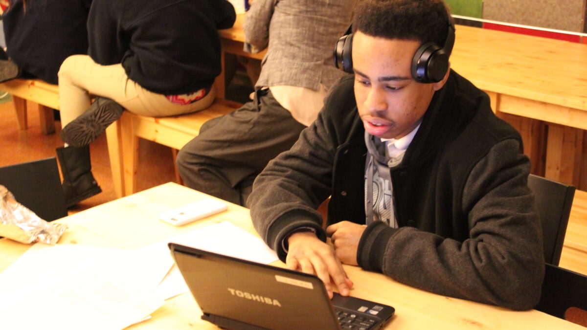 A teen wearing headphones looking at a laptop computer on a wooden table