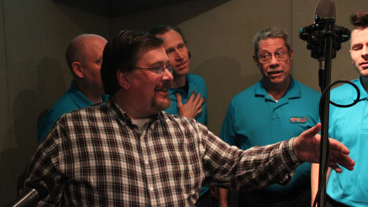 Five adults singing into a microphone in a radio studio