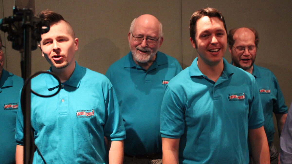 Four adults singing into a microphone in a radio studio