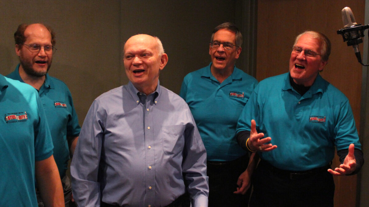 Five adults singing into a microphone in a radio studio