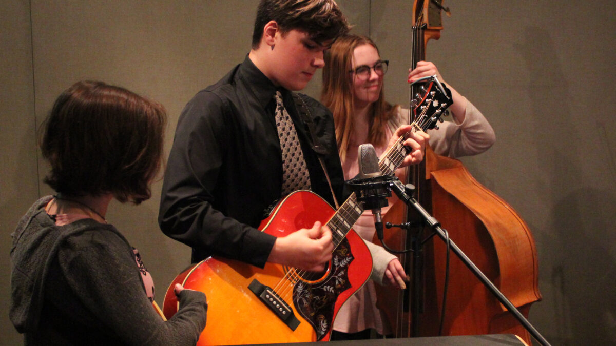 A teen playing upright bass, a teen playing acoustic guitar, and a teen standing nearby in a radio studio.