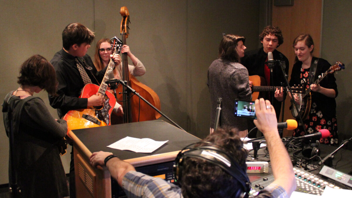 A teen playing banjo, a teen playing acoustic guitar and singing into a microphone, a teen singing into a microphone, a teen playing upright bass, a teen playing acoustic guitar, and a teen standing nearby as an adult wearing headphones sitting behind a mixing board listens in a radio studio