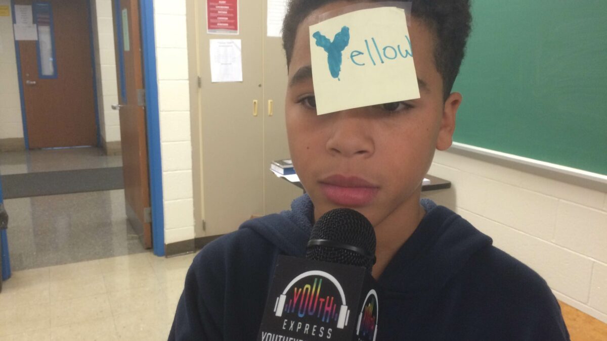 A youth wearing a post-it-note with the text "yellow" on it holding a microphone in a classroom