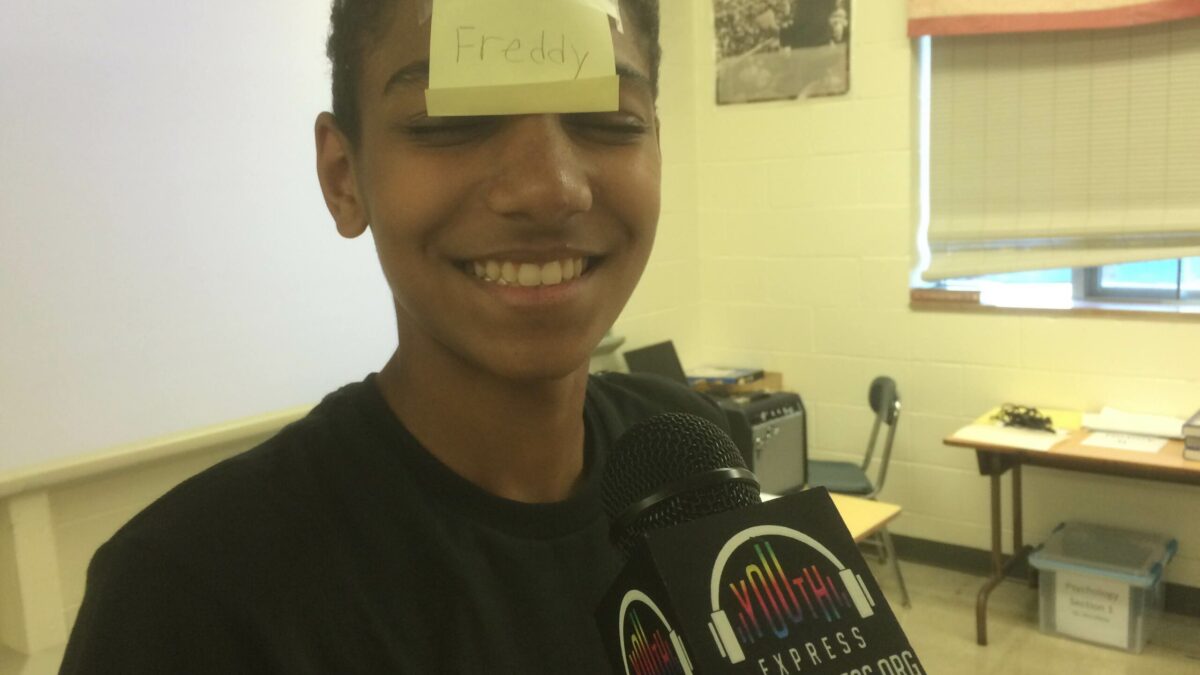 A youth wearing a post it note with the text "Freddy" on it holding a microphone in a classroom