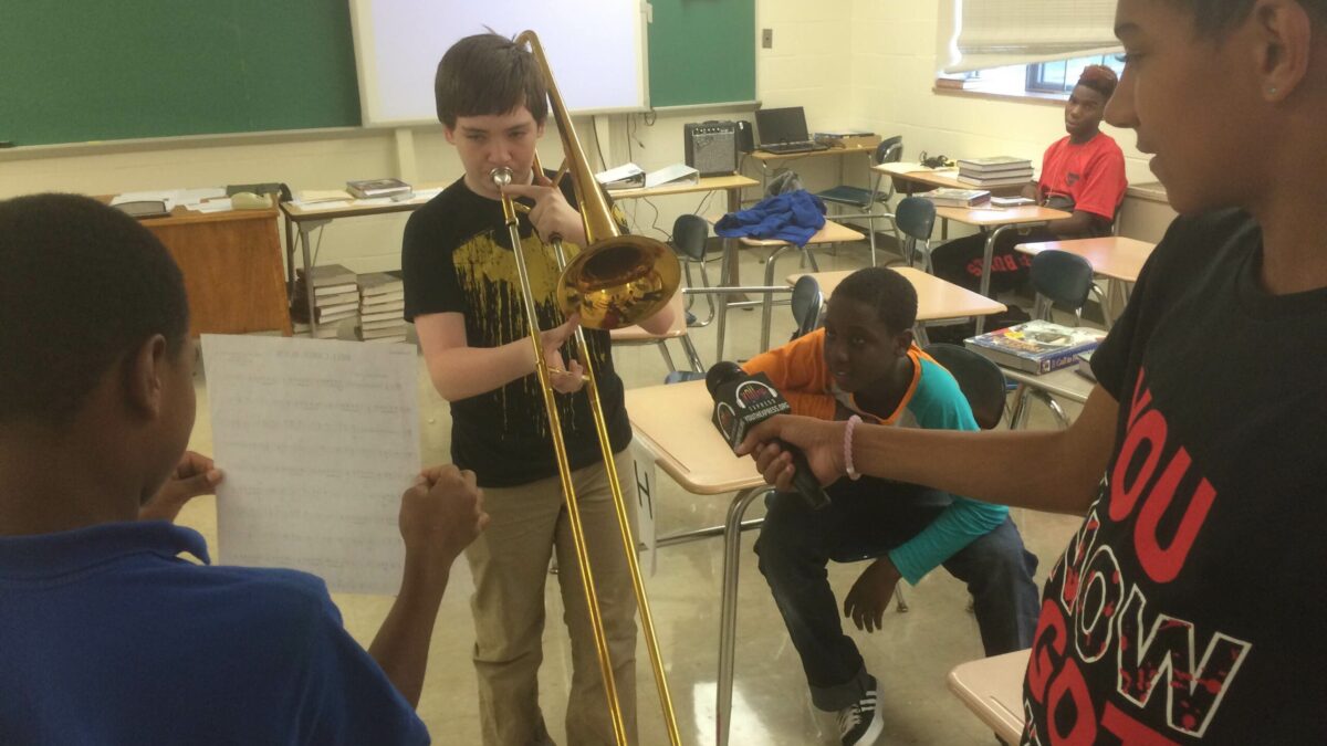 A youth holding a microphone up to another youth holding a trombone as another youth holds up a piece of paper and another youth sits at a classroom desk
