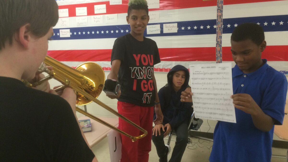 A youth holding a microphone up to another youth holding a trombone as another youth holds up a piece of paper and another youth sits at a classroom desk