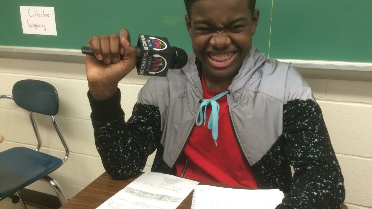 A youth smiling while holding a microphone sitting behind two pieces of paper on a classroom desk