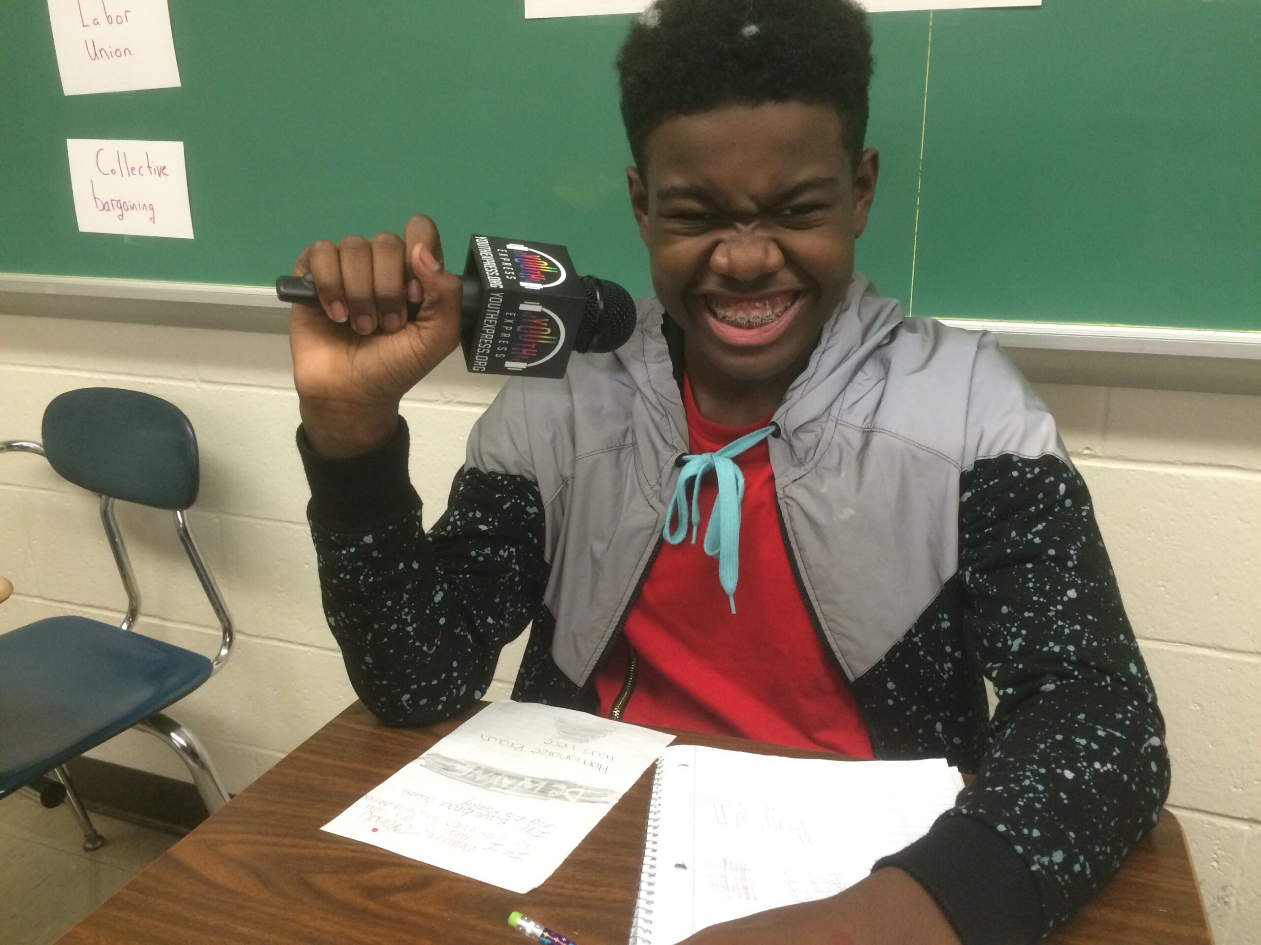 A youth smiling while holding a microphone sitting behind two pieces of paper on a classroom desk