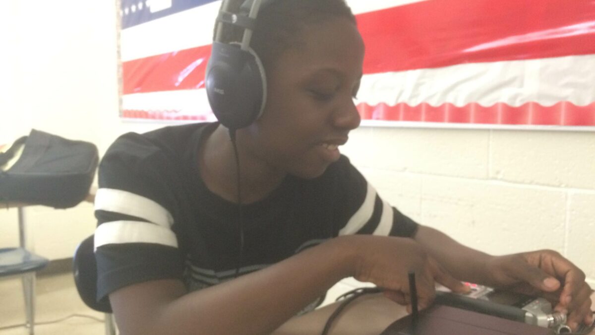 A youth smiling while wearing headphones interacting with a portable recorder sitting at a classroom desk