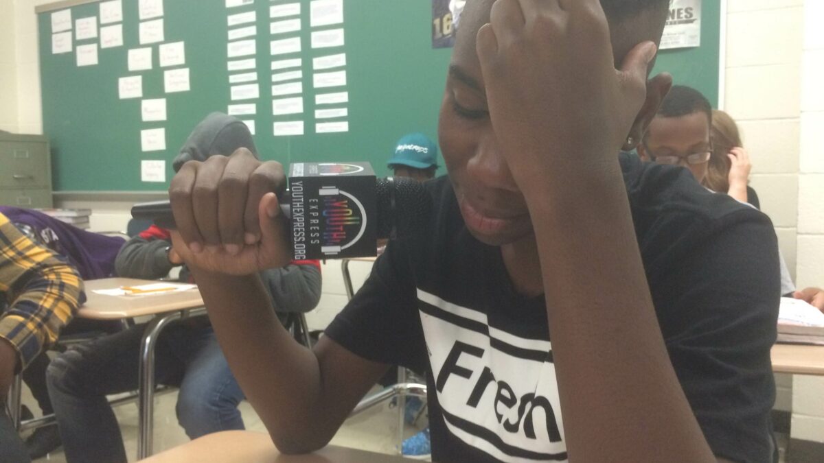A youth holding a microphone looking down at a piece of paper on a classroom desk