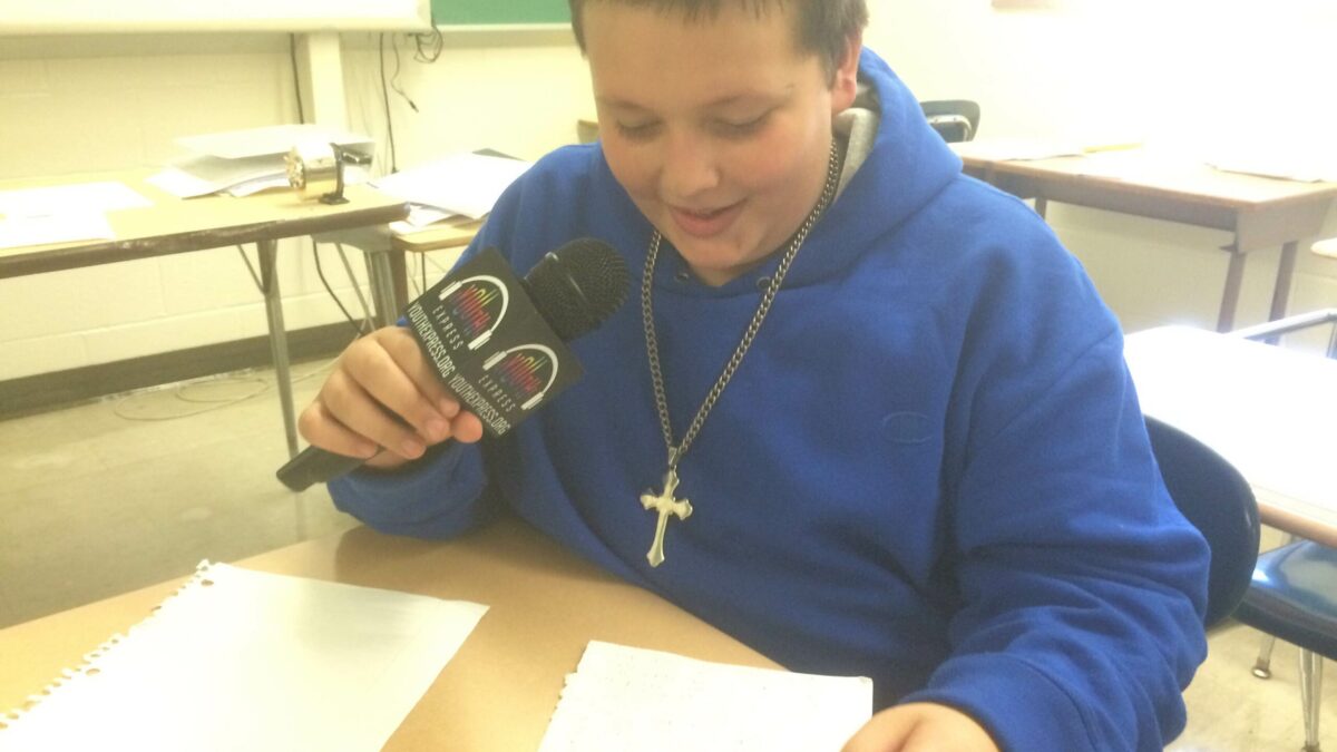 A youth holding a microphone looking down at pieces of paper on a classroom desk