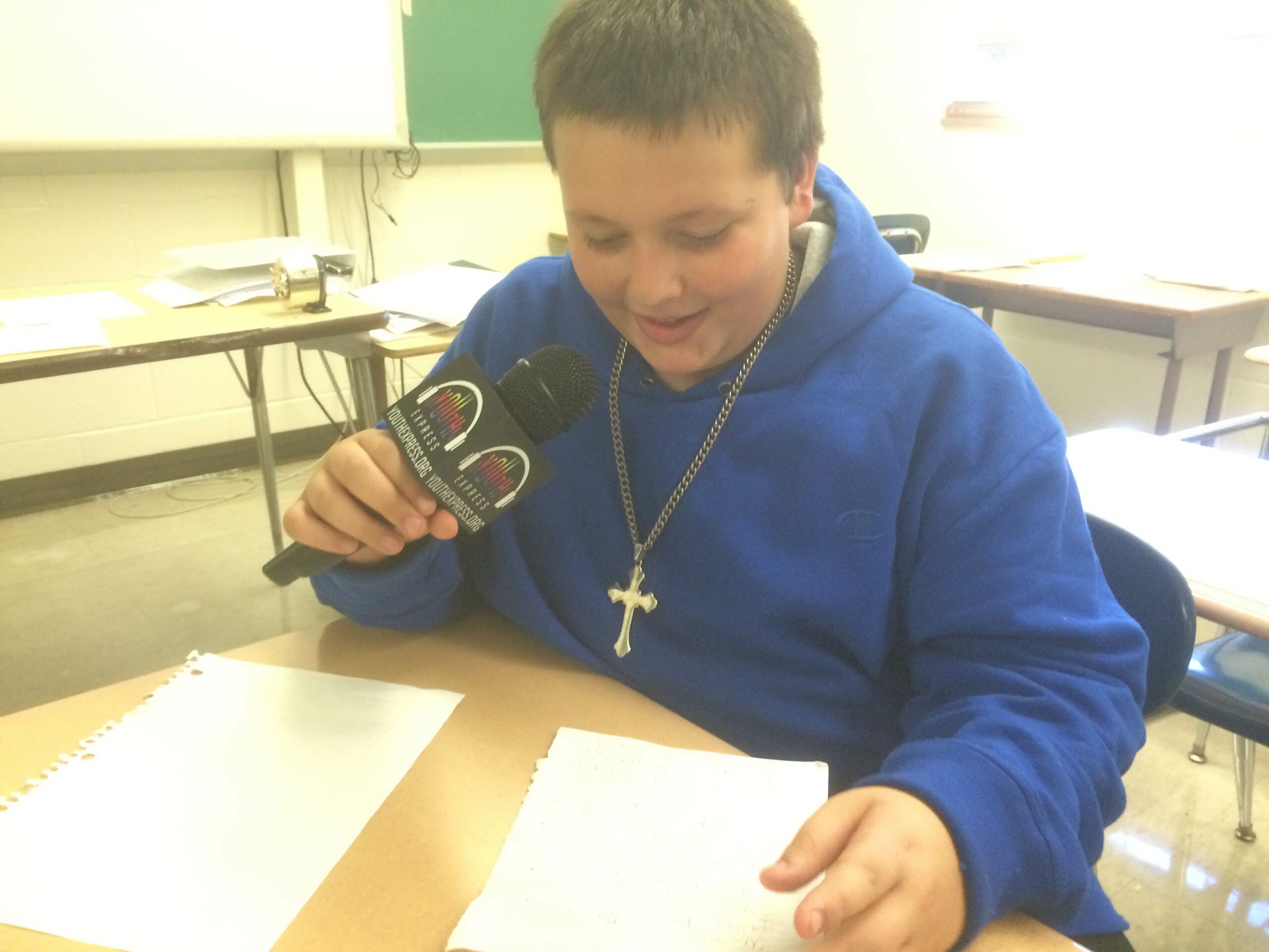 A youth holding a microphone looking down at pieces of paper on a classroom desk
