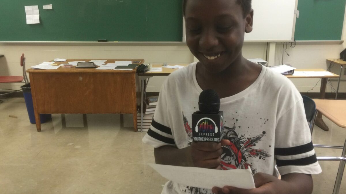 A youth smiling holding a microphone and a piece of paper in a classroom