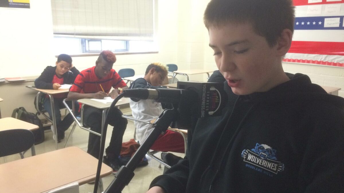 A youth speaking into a microphone and holding a piece of paper as three youth listen in the background in a classroom