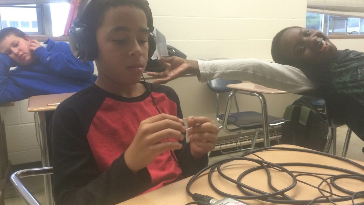 A youth wearing headphones connected to a portable recorder sitting in a classroom