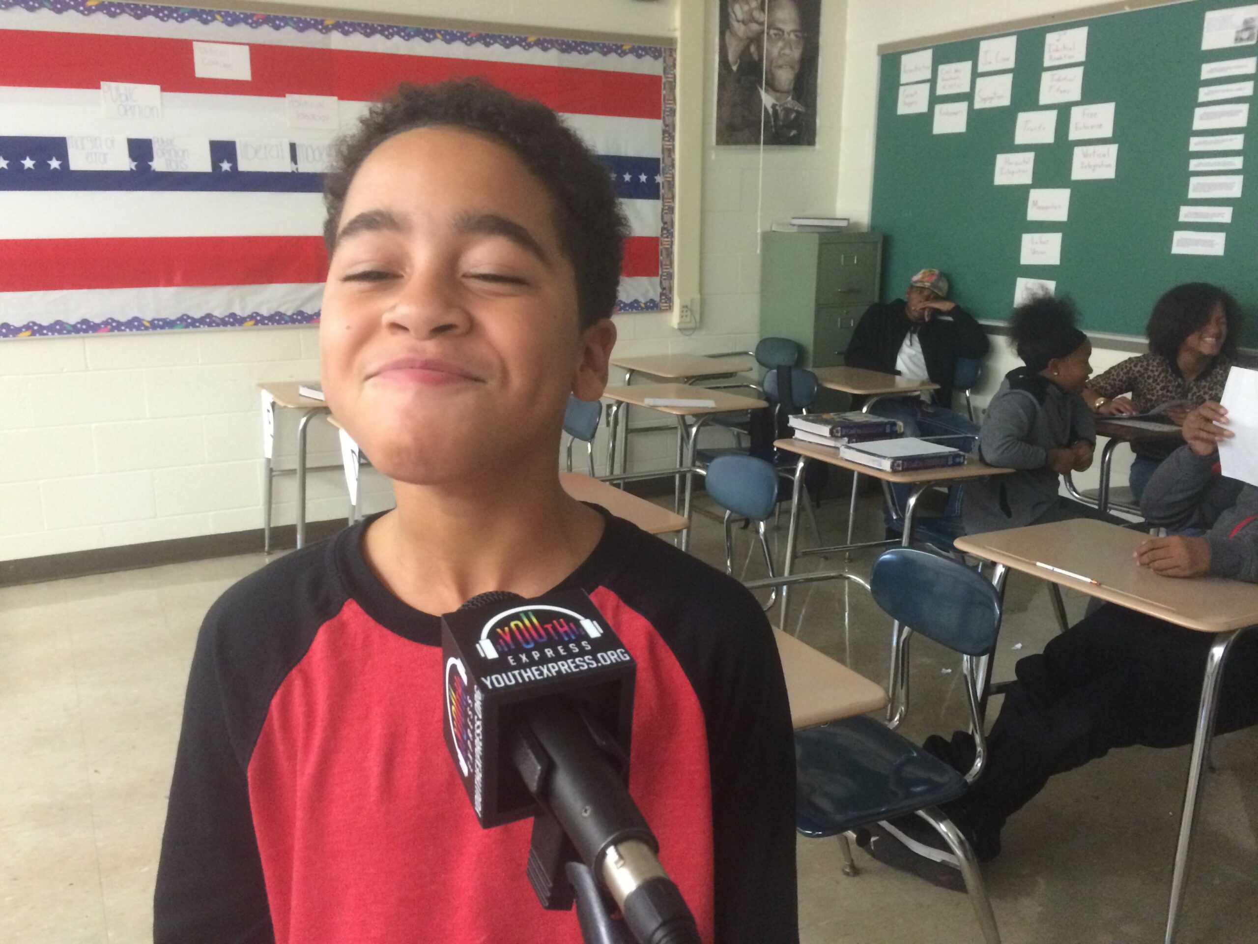 A youth smiling behind a microphone as three youth sit in the background in a classroom