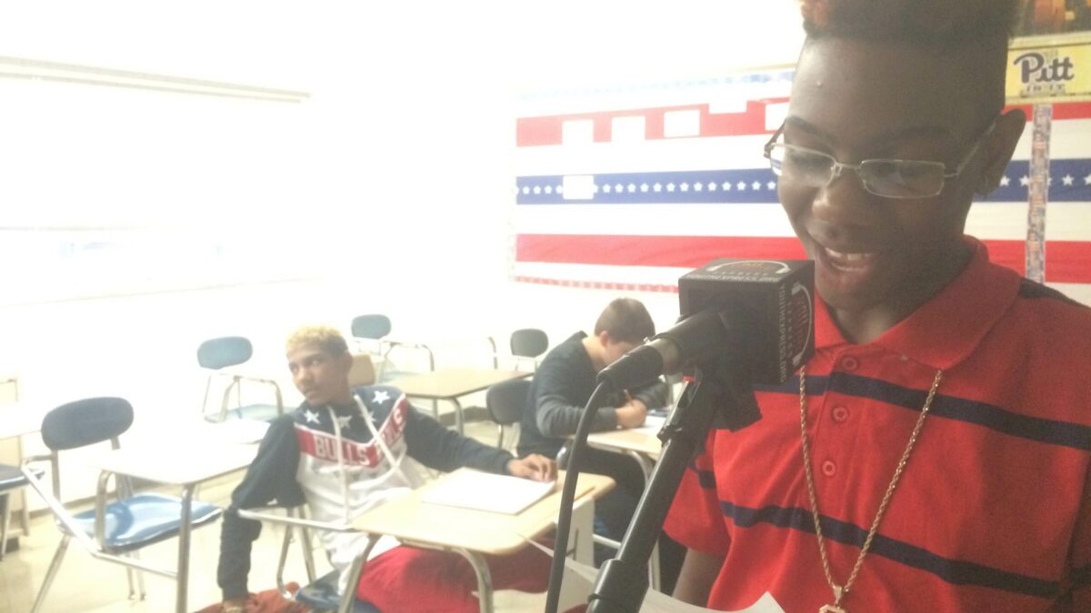 A youth holding a piece of paper speaking into a microphone as two youth listen in the background in a classroom
